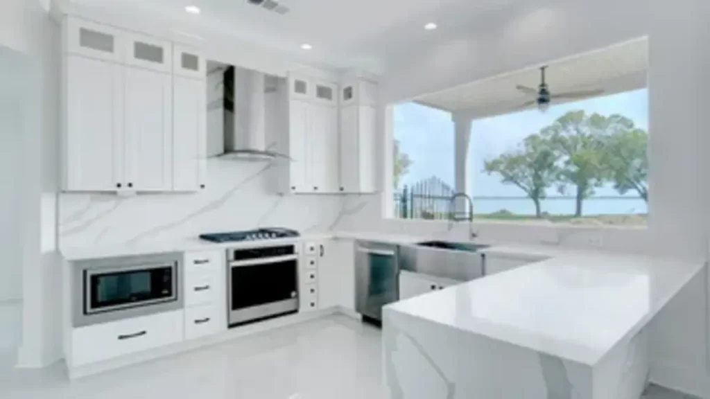 Modern white kitchen interior featuring sleek white cabinets, stainless steel appliances, and marble-look quartz countertops with a large window overlooking an outdoor view.